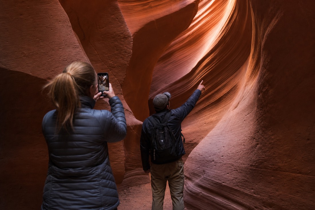Guide gesturing toward a light-filled curve of slot canyon wall while a visitor raises a phone