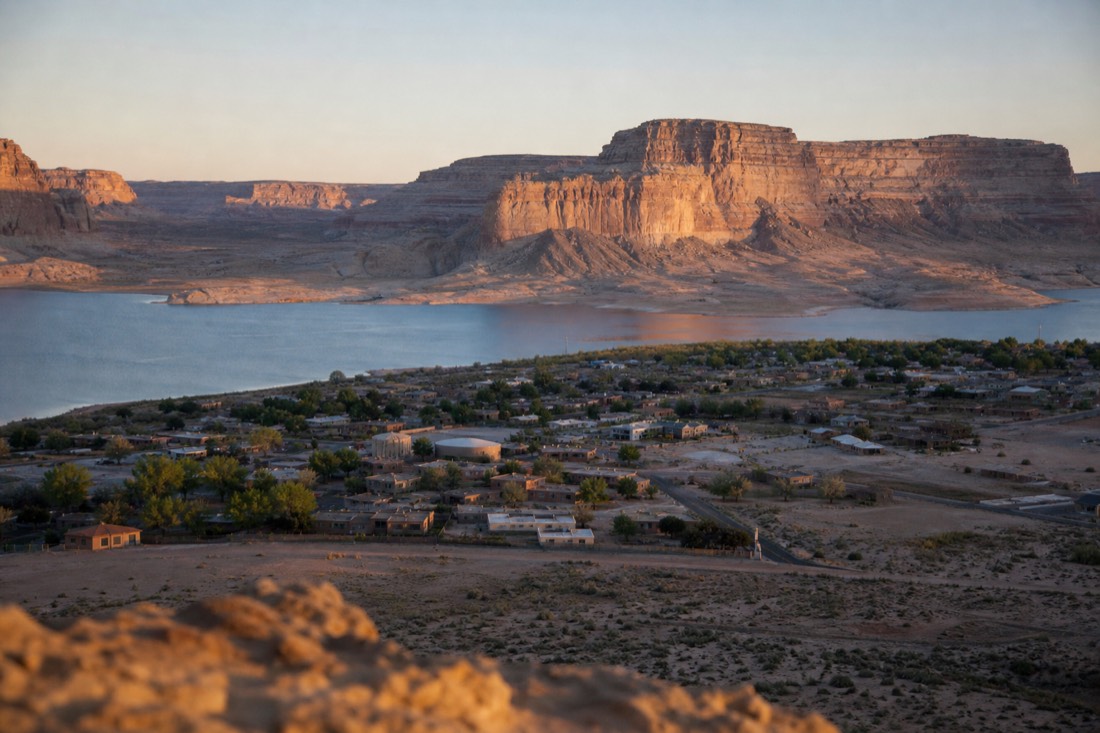 Compact desert town beside blue reservoir water with sandstone cliffs at dawn