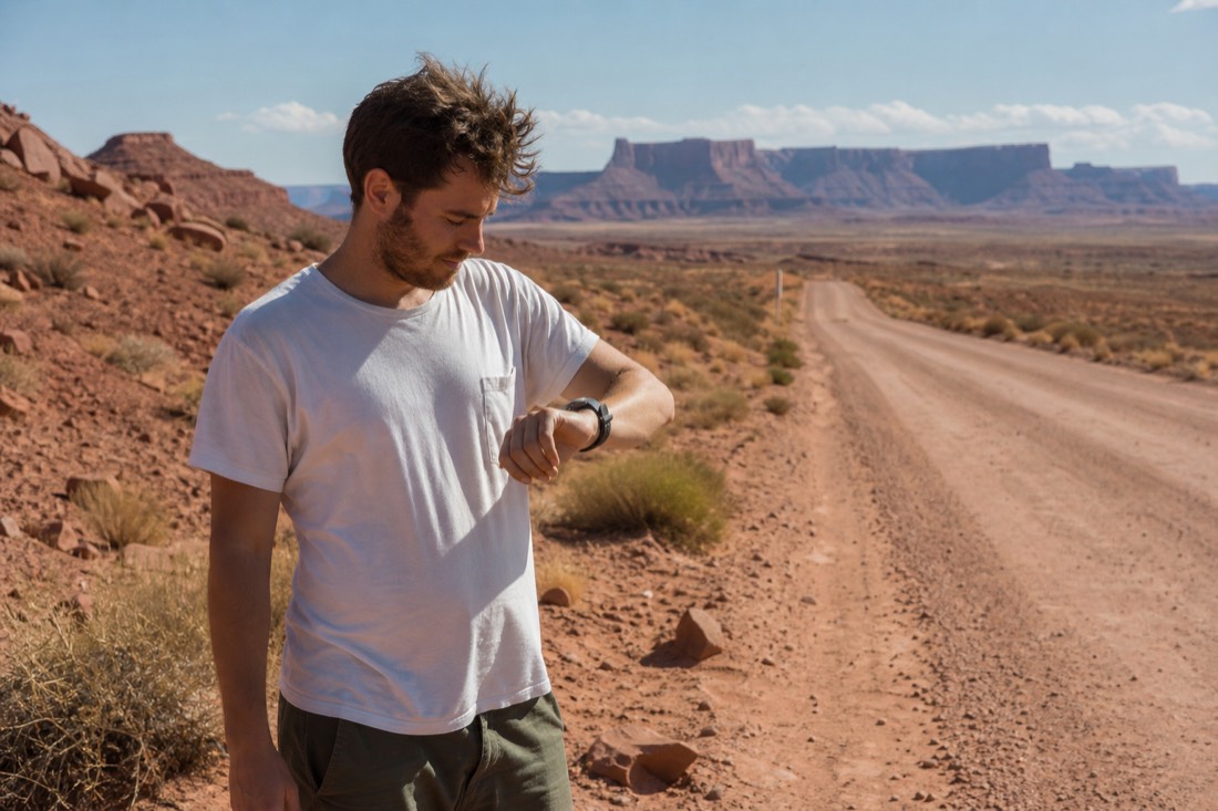 Traveler checking a wristwatch beside a dusty highway with mesa country behind
