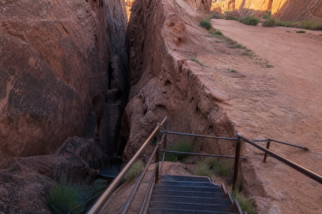 Steel stairways descending into a narrow V-shaped slot canyon beside a flat sandy canyon corridor