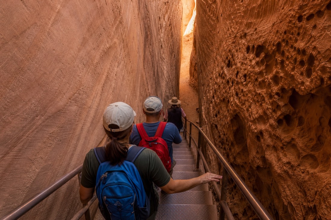 Visitors descending a metal staircase between narrow sandstone walls with a Navajo guide ahead