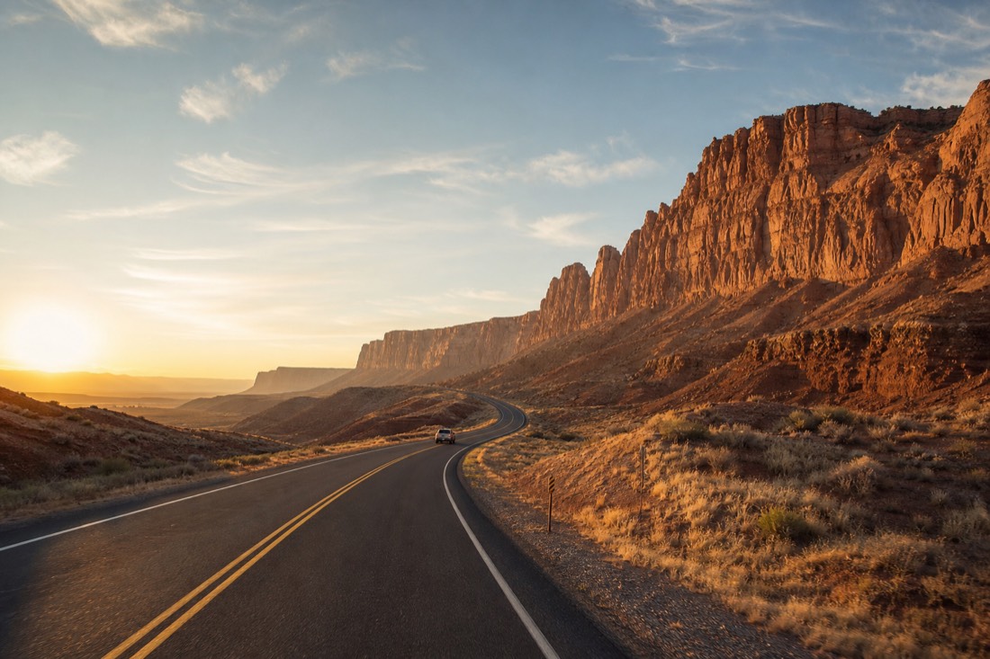 Car driving a desert highway at sunrise toward sandstone cliffs east of town